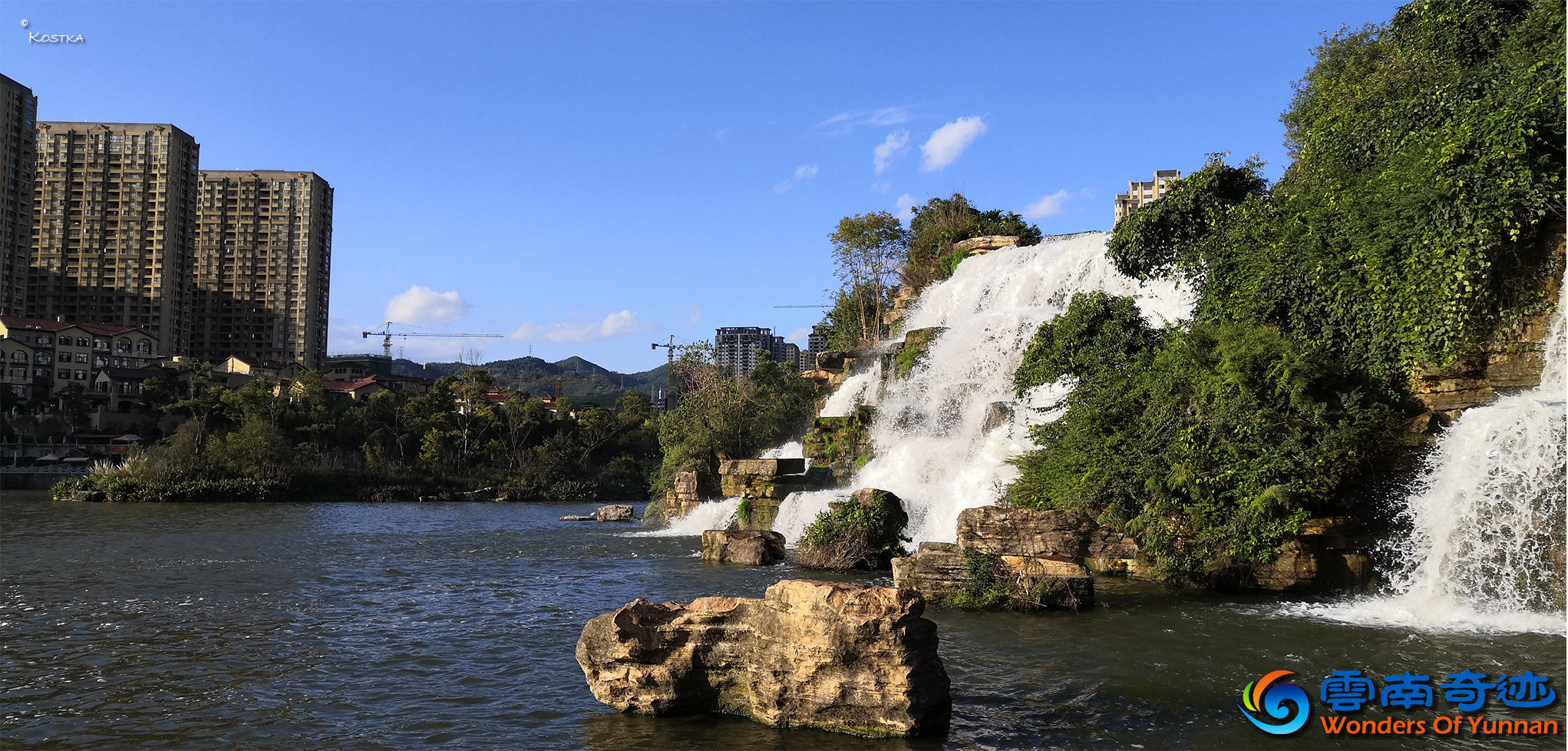 Natural waterfalls at the Waterfall Park covert with trees, plants and flowers with a view over the lower lake with a forest covert island in the middle of the lake on a sunny day